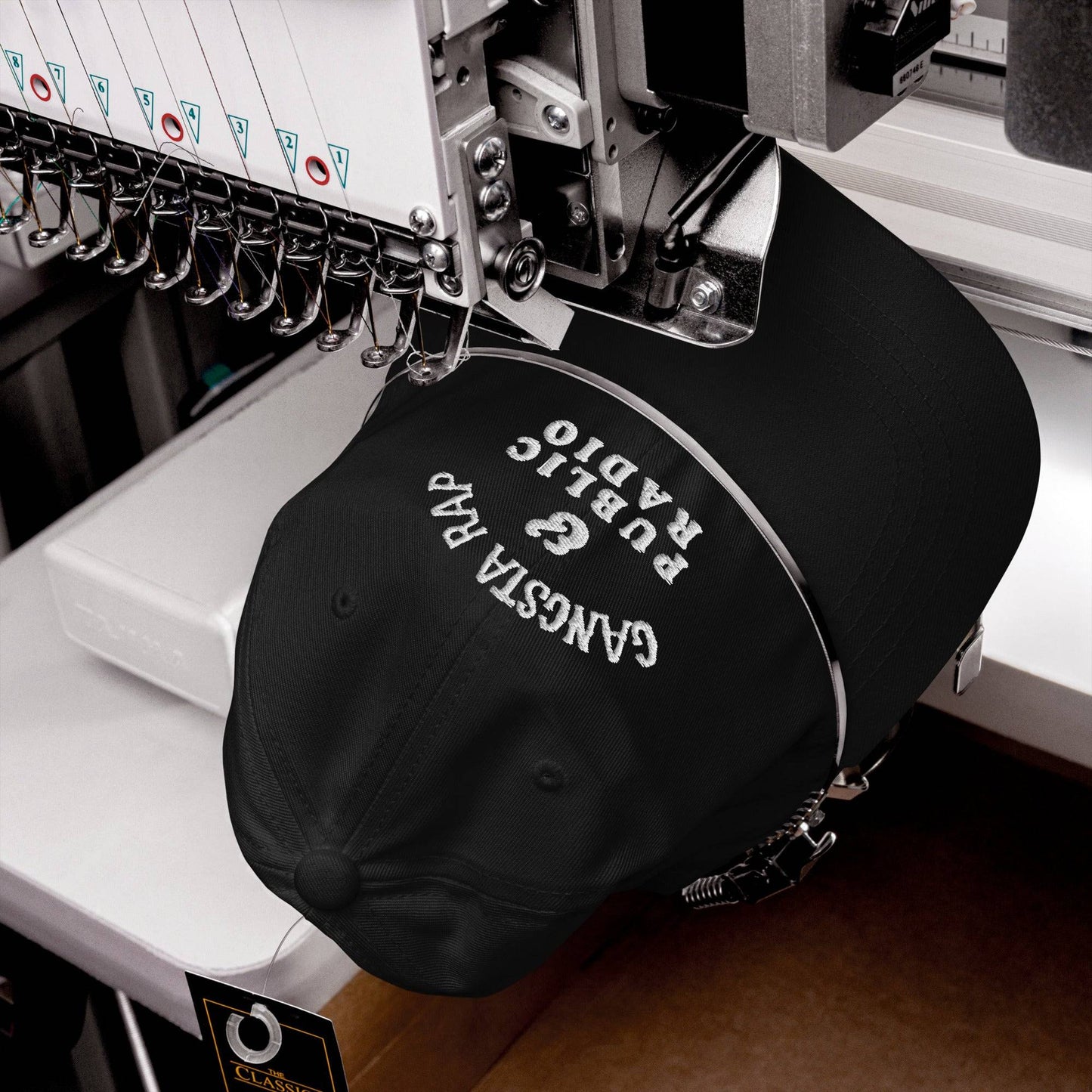 Black Gangsta Rap And Public Radio Baseball Cap being embroidered on industrial machine, showcasing USA-made craftsmanship.
