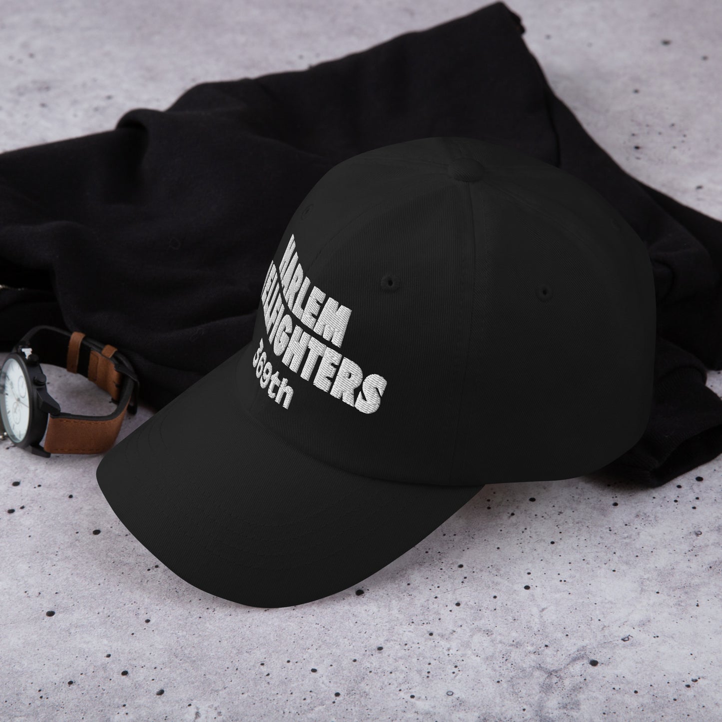 Black Harlem Hellfighters 369th cap placed on a concrete surface with watch and sweater. Stylish heritage hat for everyday wear and gifting.
