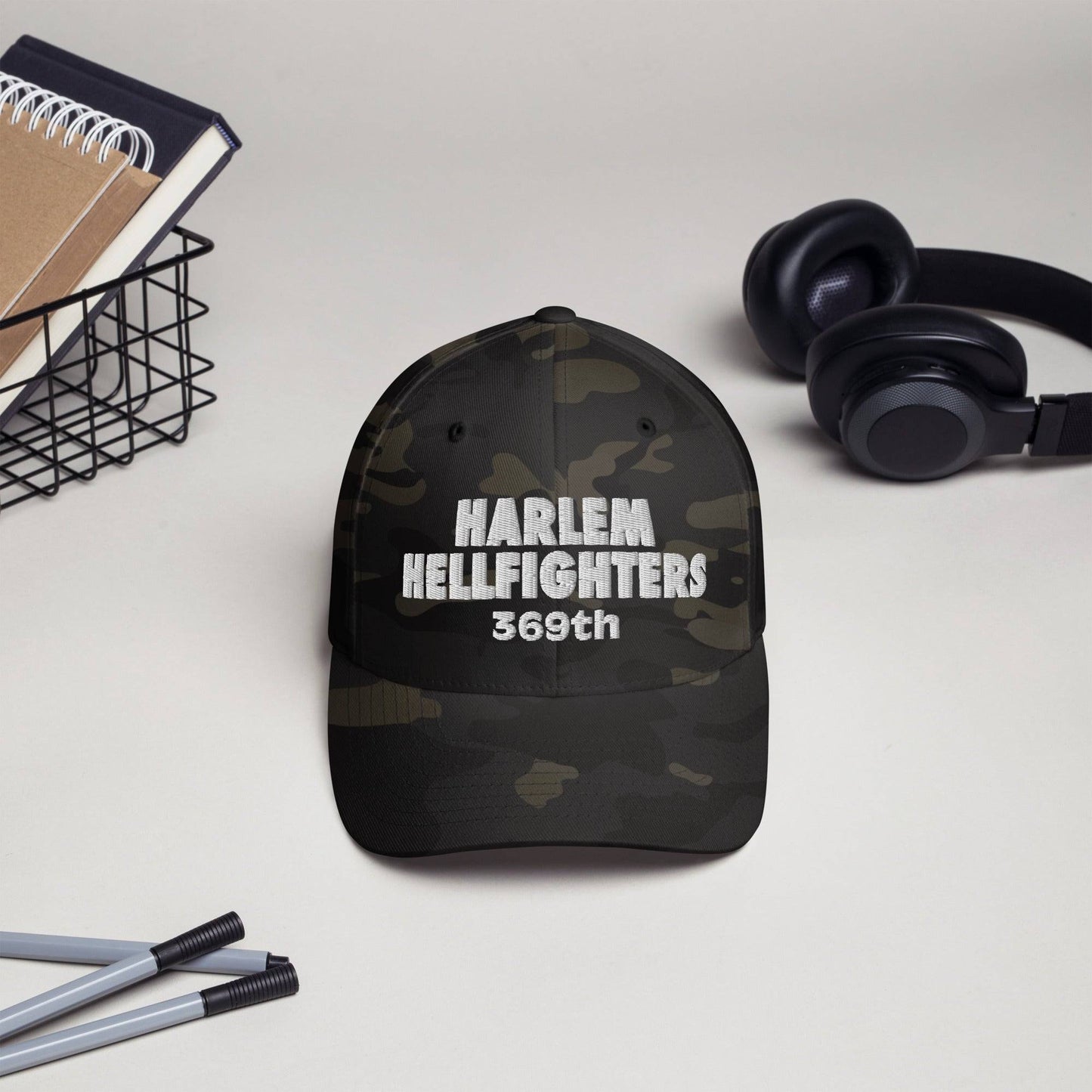 Harlem Hellfighters 369th Flexfit® Cap in black camo displayed on desk with headphones and pens. Stylish heritage hat for everyday wear.