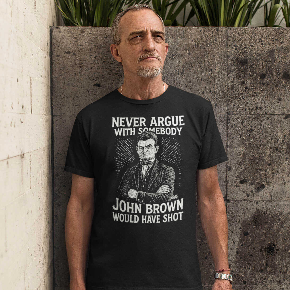 Man wearing Never Argue John Brown Would Have Shot Unisex Staple T-Shirt in black, standing confidently against a concrete wall.
