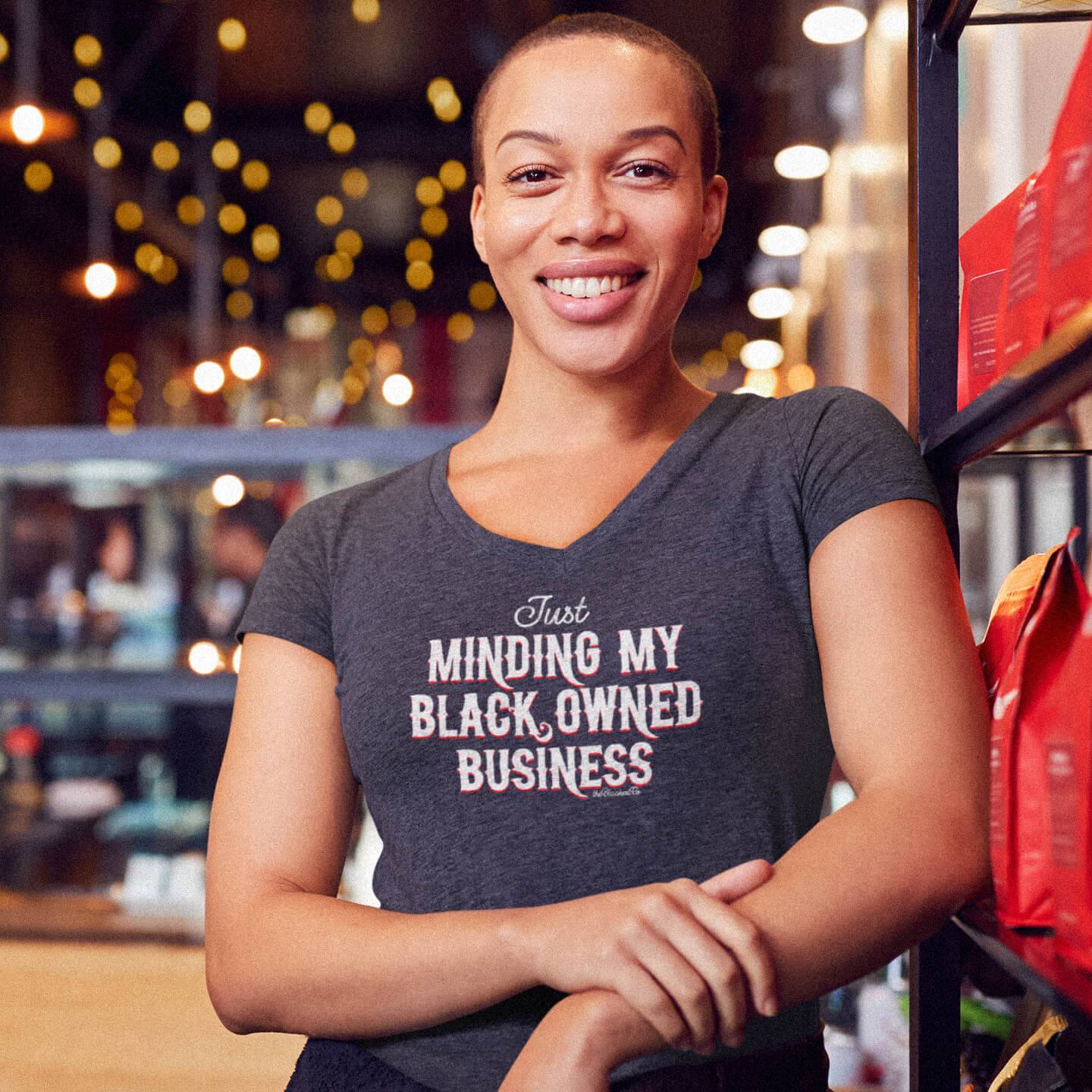 Smiling woman wearing charcoal Minding My Black Owned Business Womens V-neck T-shirt in a cozy café, showcasing stylish empowerment.