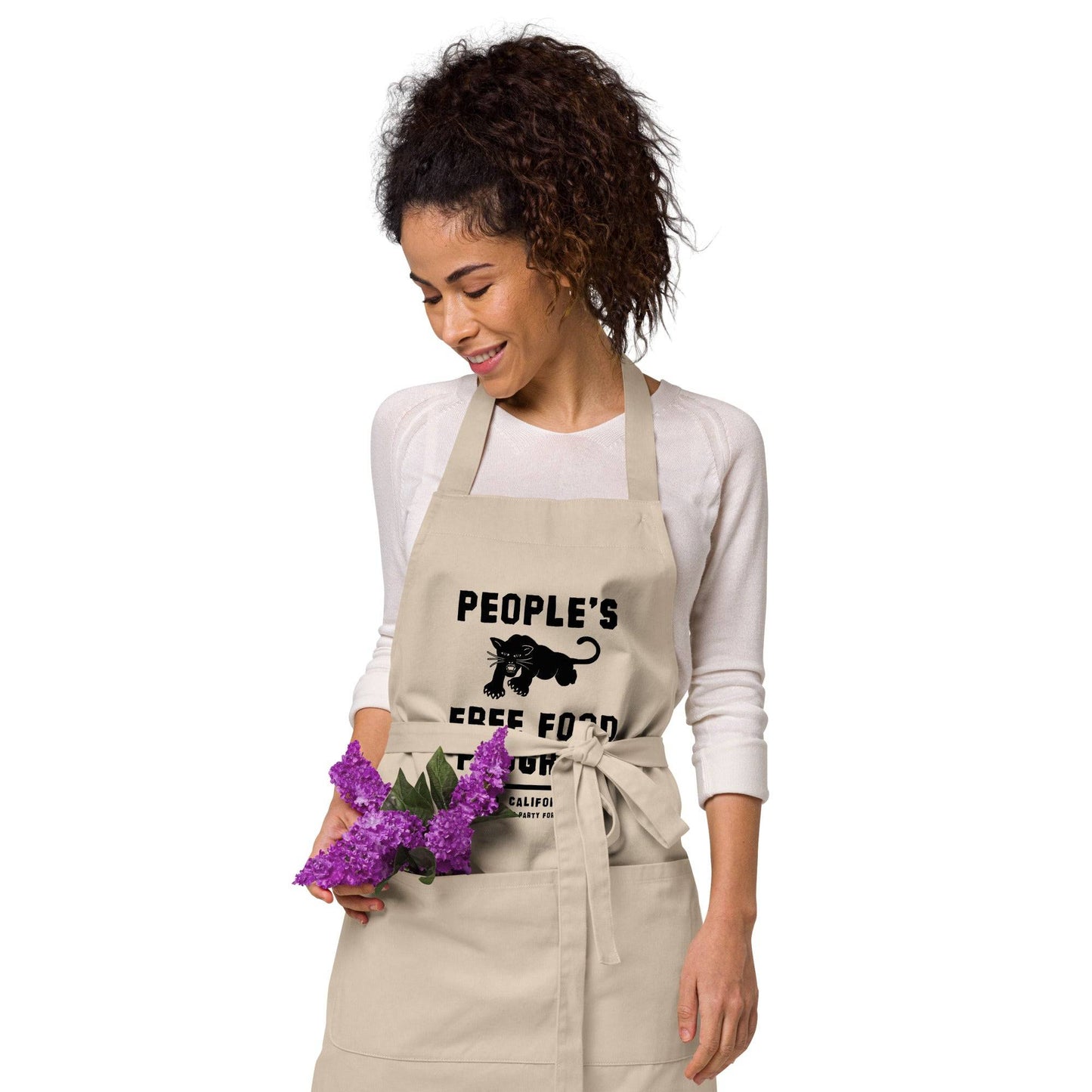 Woman holding purple flowers and smiling in beige BPP People's Free Food Program Organic Cotton Apron with waist tie and bold front print.