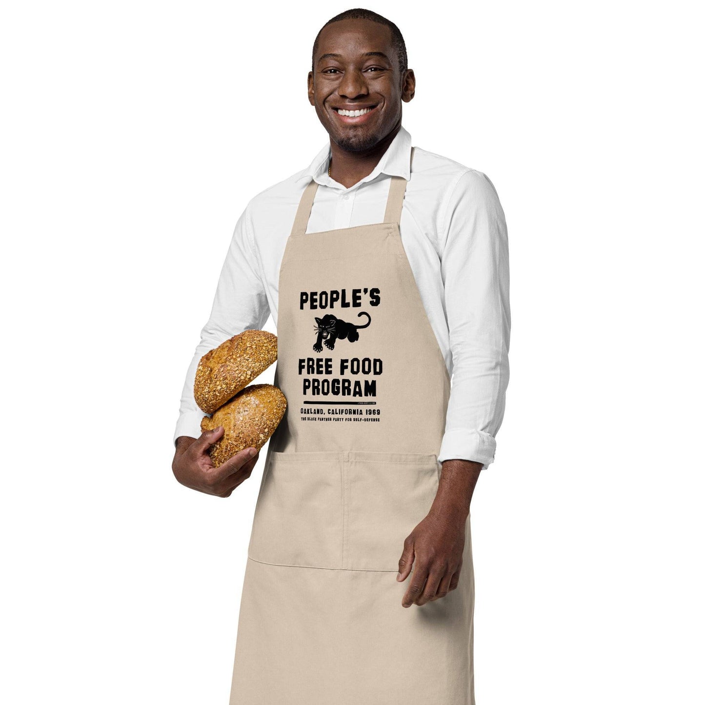 Man holding fresh bread while wearing BPP People's Free Food Program Organic Cotton Apron in beige, celebrating Black Panther legacy.