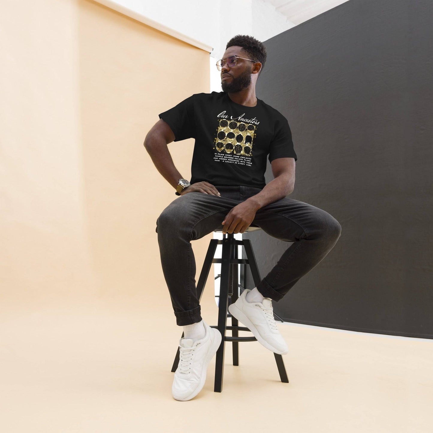 Stylish man seated in black Our Ancestors Braided Escape Routes Staple T-Shirt, showcasing legacy of hidden braid messages.