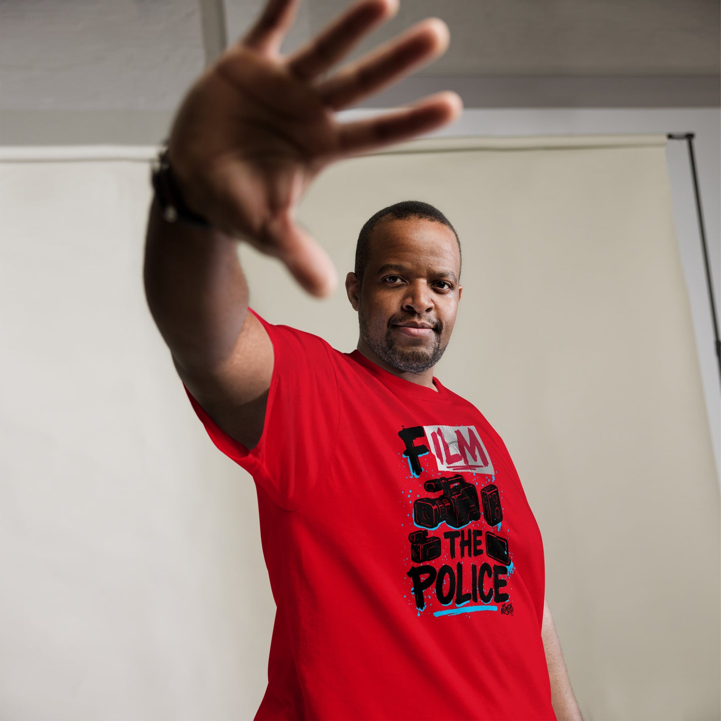 Man wearing red Film The Police Staple T-shirt by TheBlackest Co., showcasing bold activist design in a casual indoor setting.