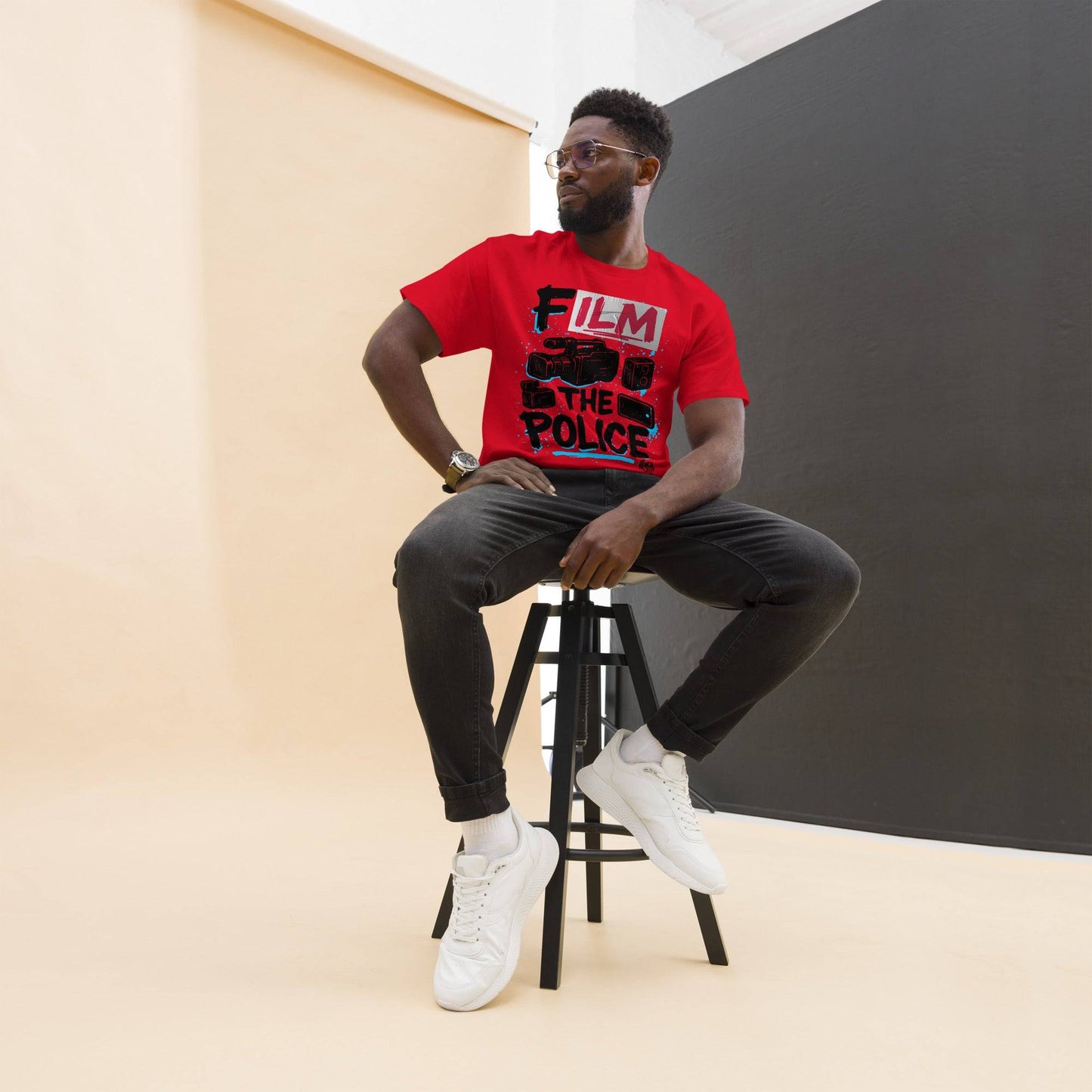 Model seated on stool wearing red Film The Police Staple T-shirt by TheBlackest Co., styled with black jeans and white sneakers.