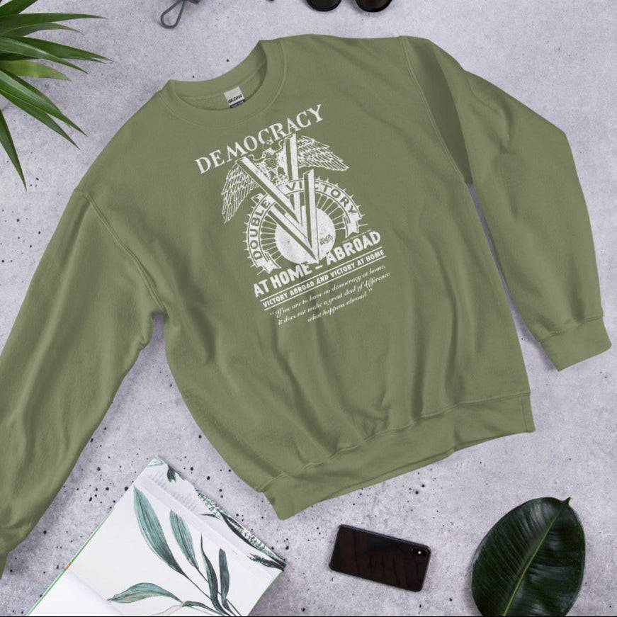 Flat lay of olive green Double V Campaign WWII Black Soldiers Sweatshirt with historical print, styled with plants and a book for lifestyle appeal.
