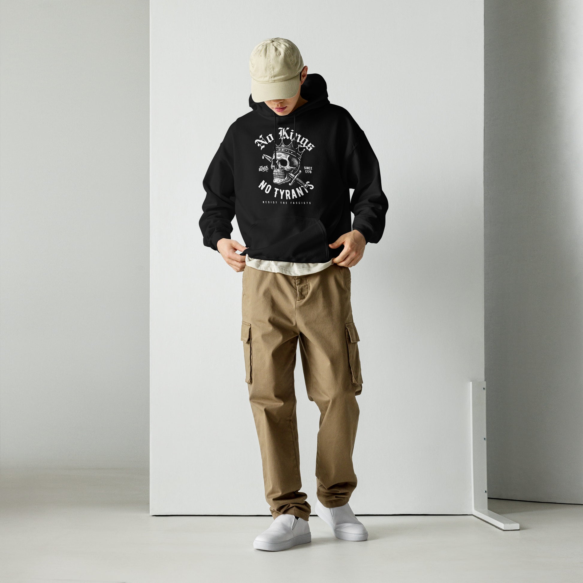 Man wearing No Kings No Tyrants Skull Protest Hoodie by TheBlackest Co., styled with khaki cargo pants and white sneakers in a modern studio setting.