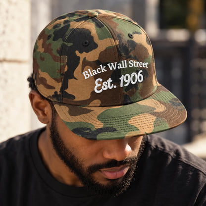 A man wearing a camouflage baseball cap with 'Black Wall Street Est. 1906' embroidered in white on the front, photographed outdoors with a blurred stone wall and railing in the background.