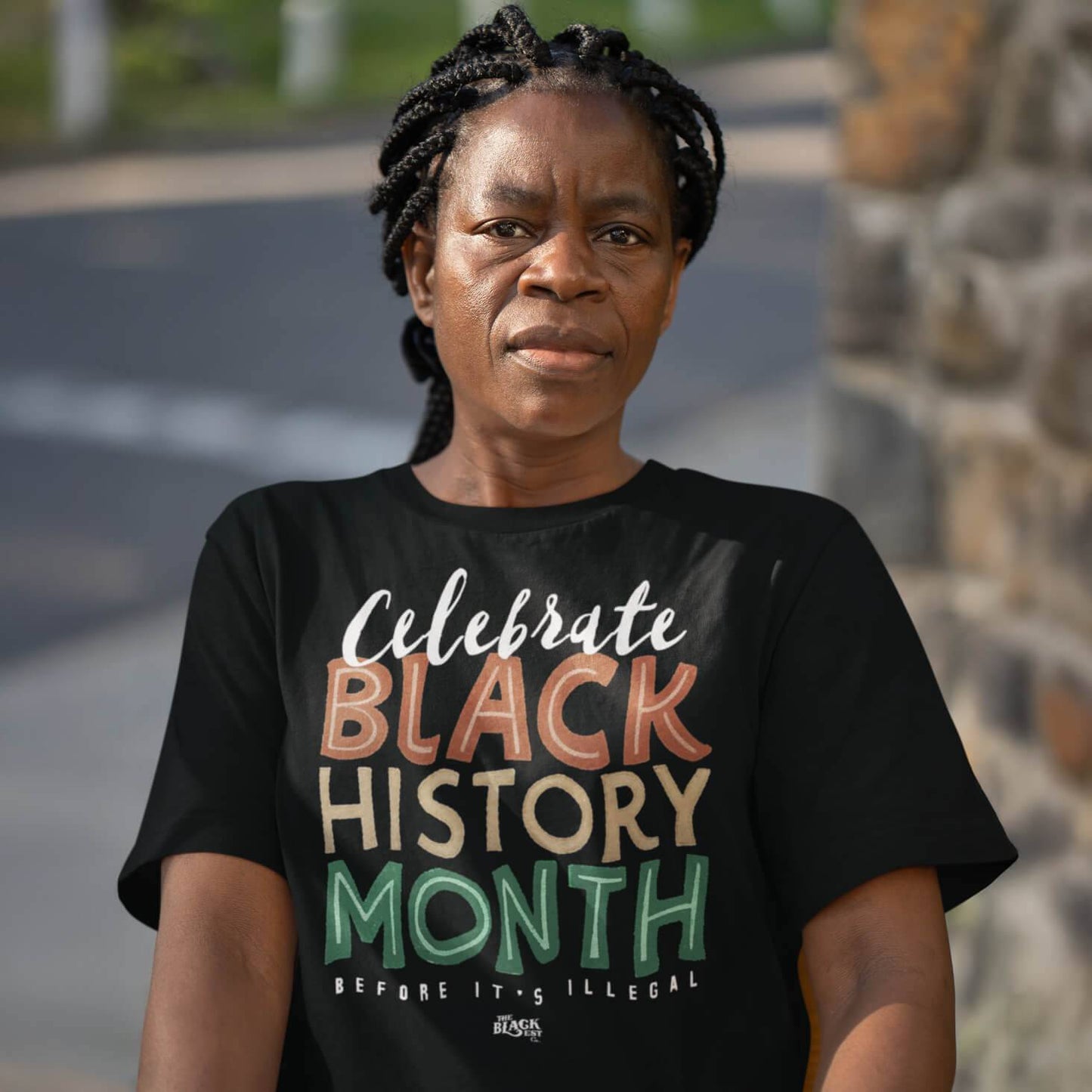 Woman wearing a black 'Celebrate Black History Month Before It's Illegal' t-shirt outdoors. Stylish unisex graphic tee for Black History Month.