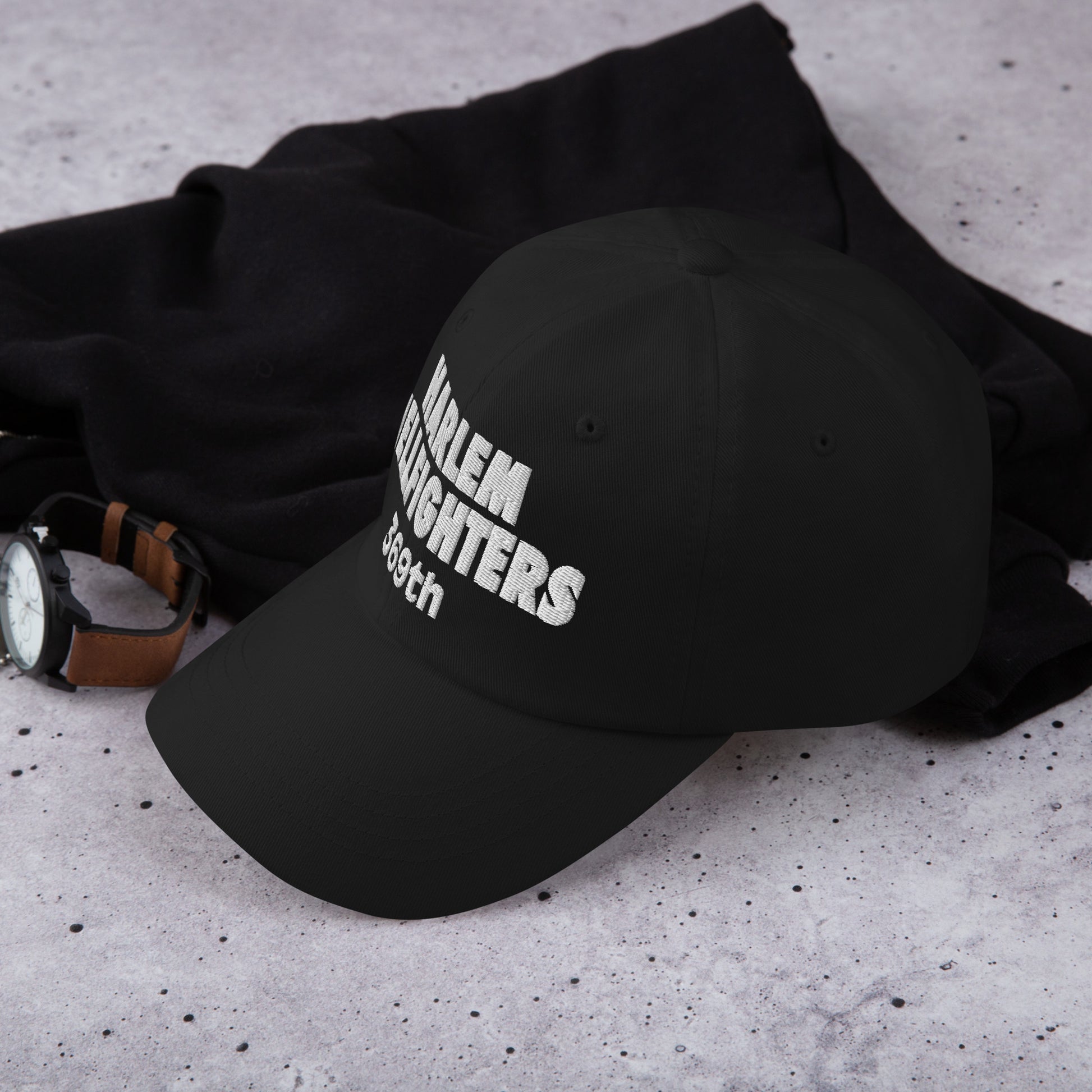 Black Harlem Hellfighters 369th cap placed on a concrete surface with watch and sweater. Stylish heritage hat for everyday wear and gifting.