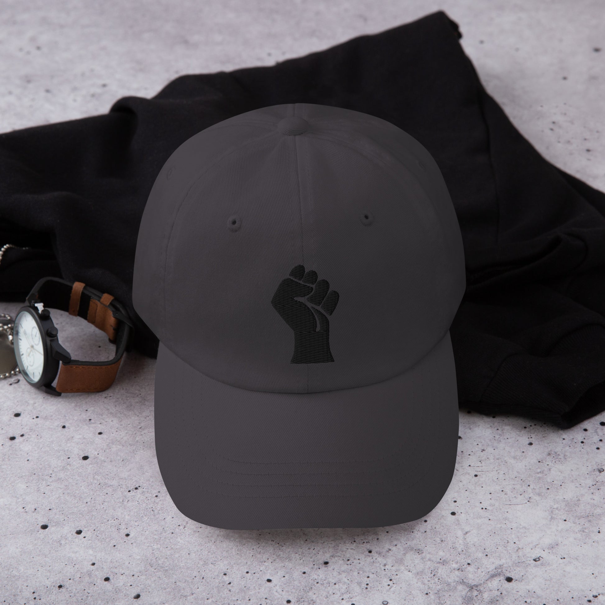 Black On Black Power Fist Embroidered Baseball Cap laid flat beside accessories, highlighting its everyday wear appeal and activism message.
