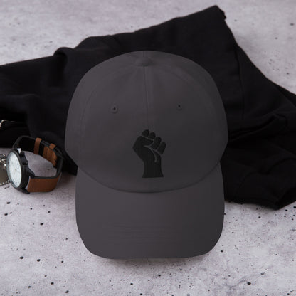Black On Black Power Fist Embroidered Baseball Cap laid flat beside accessories, highlighting its everyday wear appeal and activism message.