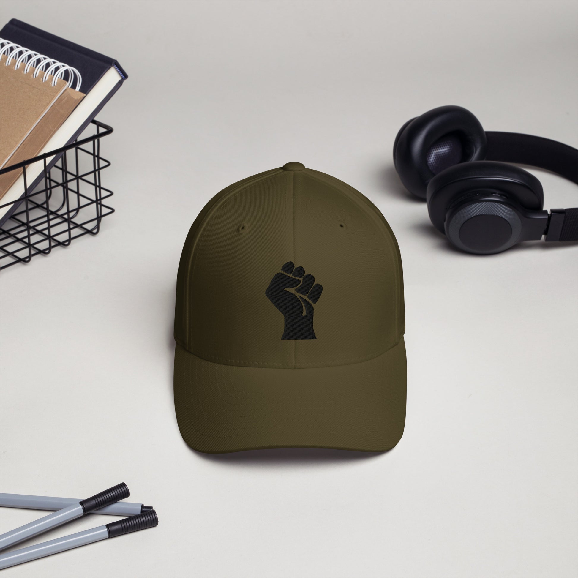 Olive green Flexfit structured cap with black embroidered power fist by TheBlackest Co., shown on a desk with headphones and pens.