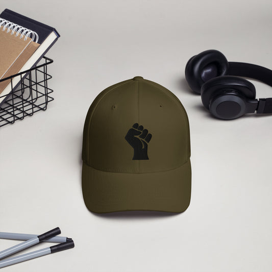 Olive green Flexfit structured cap with black embroidered power fist by TheBlackest Co., shown on a desk with headphones and pens.