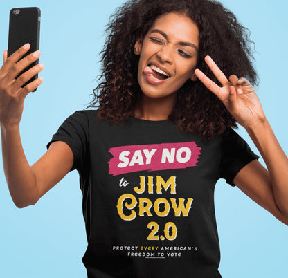A woman with curly hair takes a selfie while wearing a black graphic t-shirt that reads 'SAY NO to JIM CROW 2.0' in pink and yellow text, set against a solid light blue background.