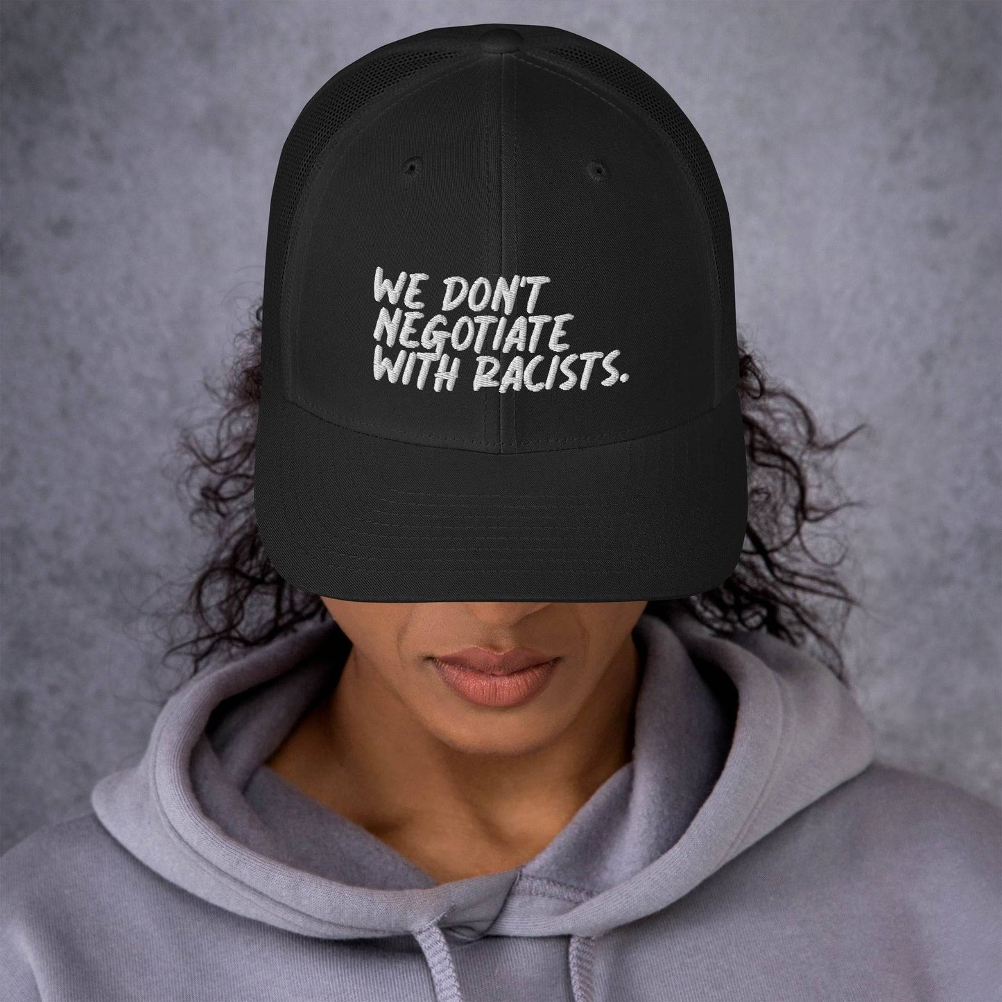front view of smiling woman wearing black trucker hat with text that reads we don't negotiate with racists embroidered