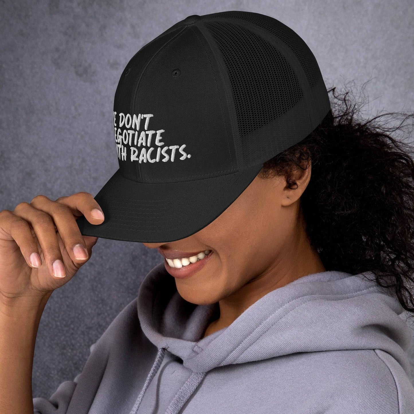 side view of smiling woman wearing black trucker hat with text that reads we don't negotiate with racists embroidered