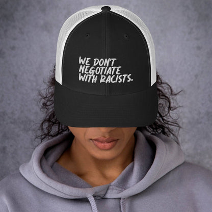 front view of smiling woman wearing white and black trucker hat with text that reads we don't negotiate with racists embroidered