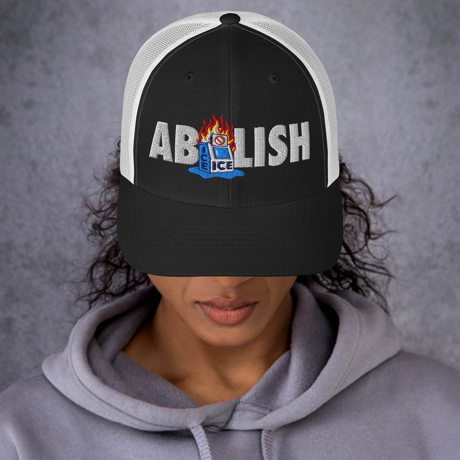 Black ABOLISH Burning ICE Machine Trucker Hat by TheBlackest Co. worn by woman, featuring embroidered statement and curved visor fit.