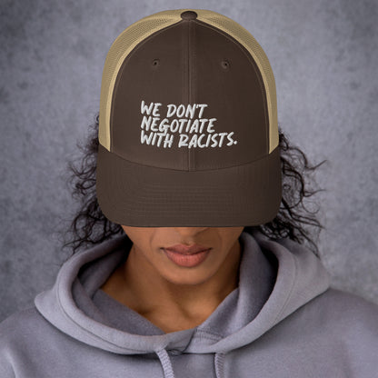 front view of smiling woman wearing brown and tan trucker hat with text that reads we don't negotiate with racists embroidered