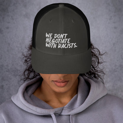 front view of smiling woman wearing grey and black trucker hat with text that reads we don't negotiate with racists embroidered