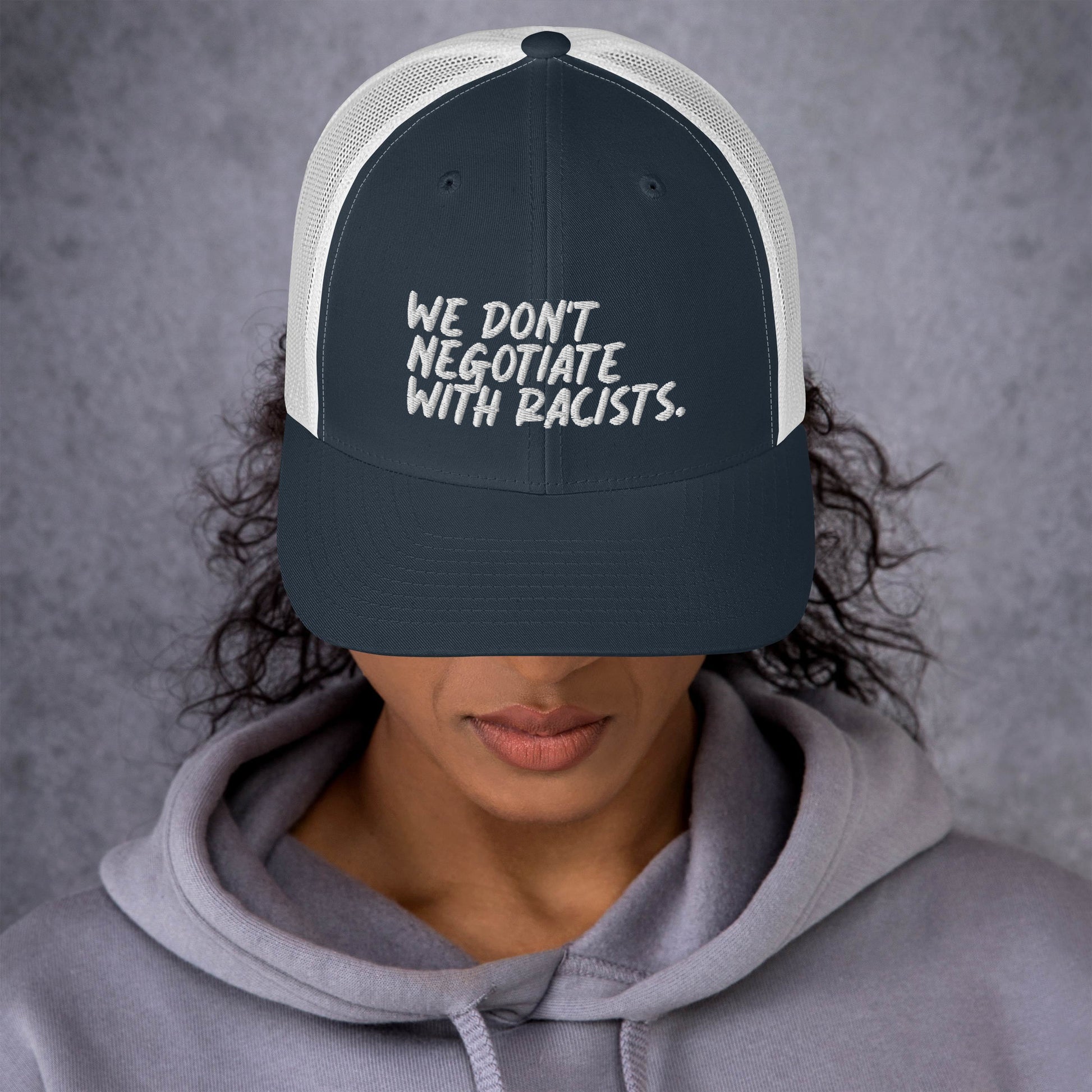 front view of smiling woman wearing white and blue trucker hat with text that reads we don't negotiate with racists embroidered