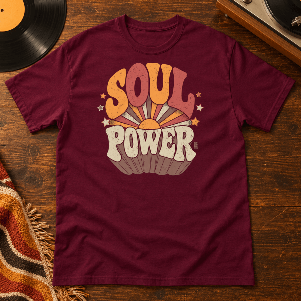 Maroon t-shirt with "Soul Power" in retro 70s lettering over an orange and red sunburst, laid flat on a wood table next to a vinyl record
