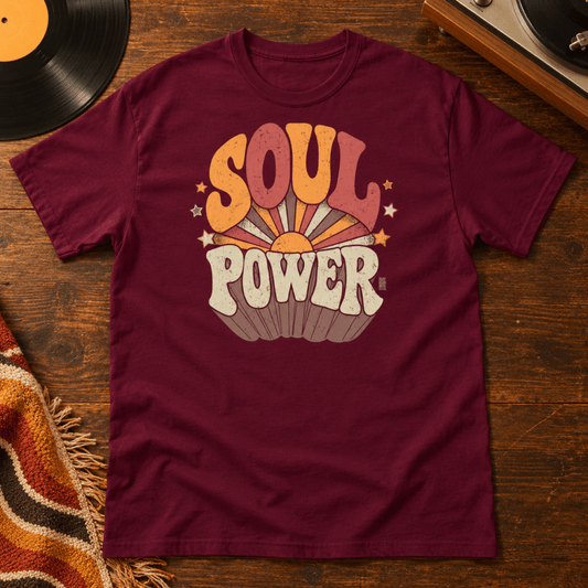 Maroon t-shirt with "Soul Power" in retro 70s lettering over an orange and red sunburst, laid flat on a wood table next to a vinyl record