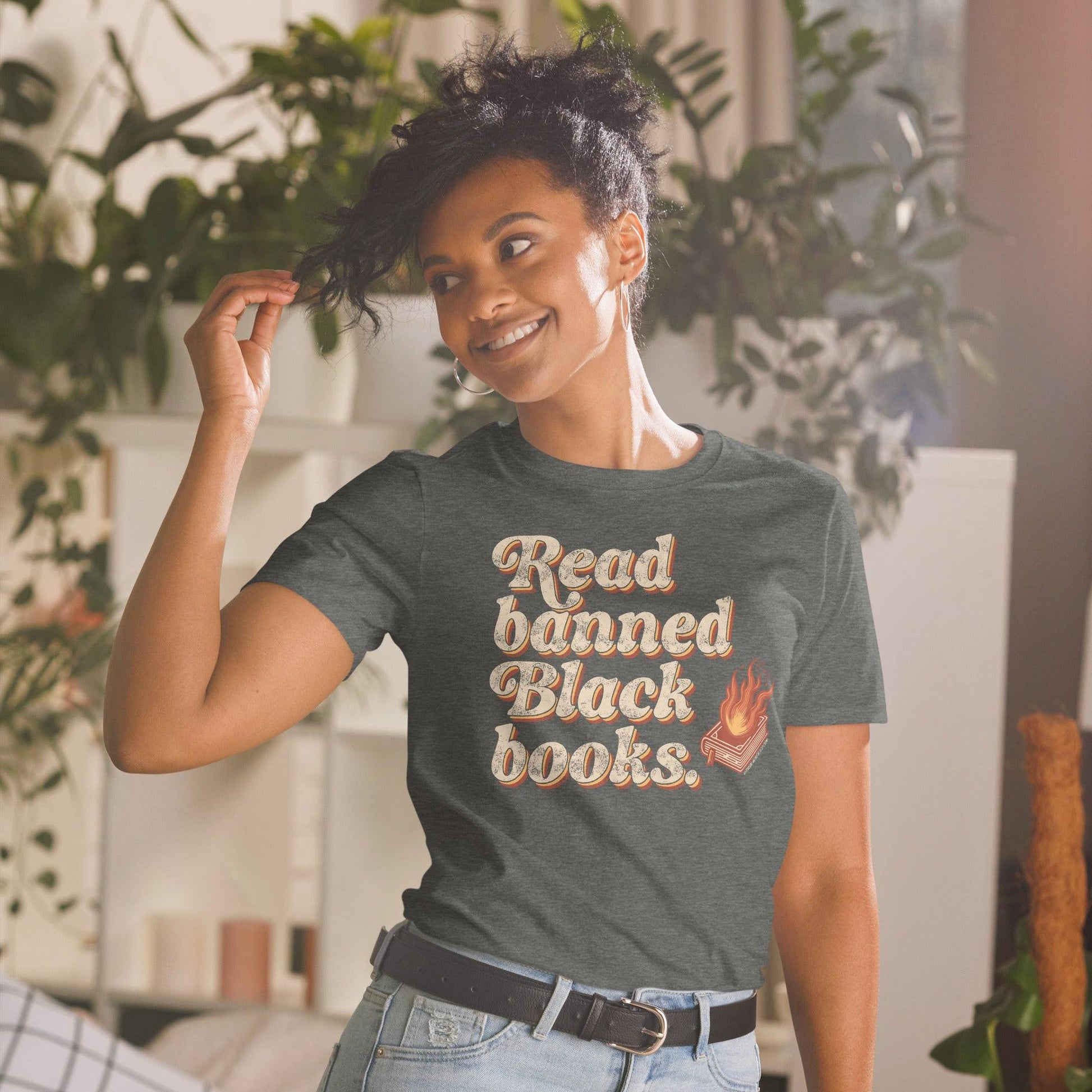 Woman wearing a dark gray 'Read Banned Black Books' t-shirt with a flaming book graphic, styled casually in a bright, cozy room.