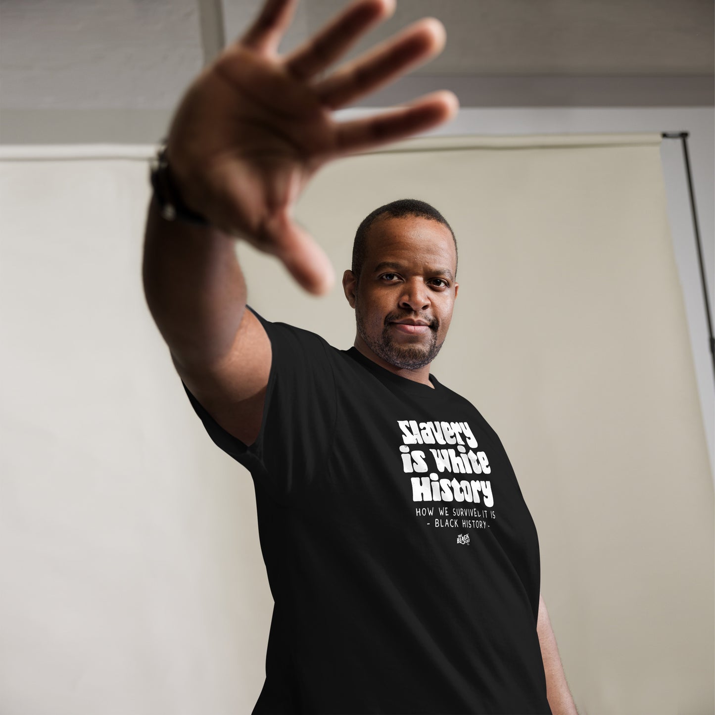 Man wearing a black unisex t-shirt with the text 'Slavery Is White History, How We Survived It Is Black History,' extending his hand forward.