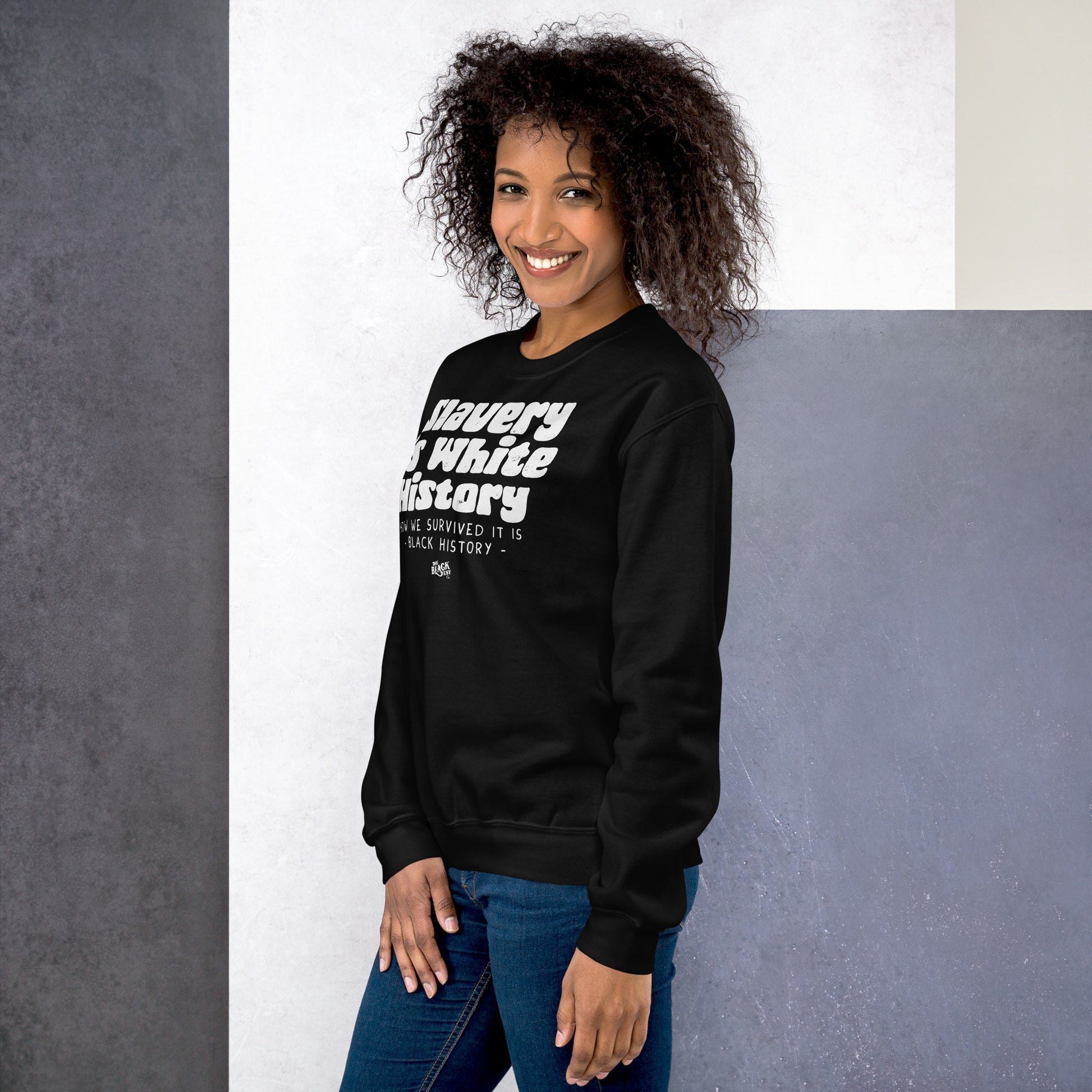 Smiling woman wearing a black 'Slavery Is White History' sweatshirt by TheBlackest Co., paired with jeans, standing against a modern backdrop.