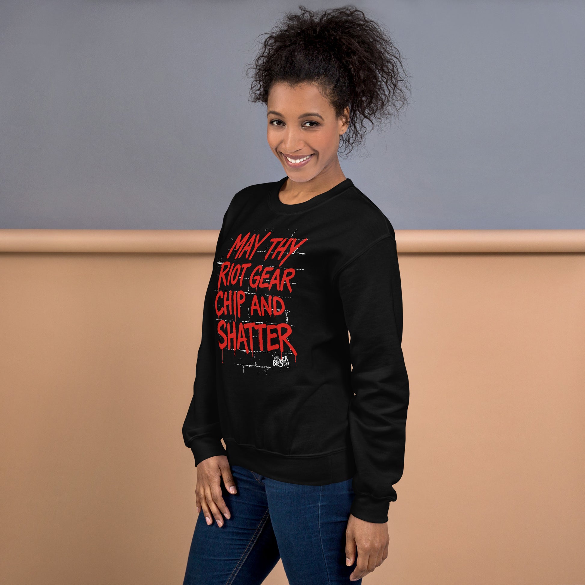 Woman wearing May Thy Riot Gear Chip And Shatter Unisex Sweatshirt in black, showcasing bold activist message in red print.