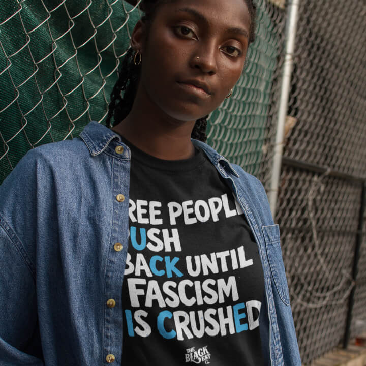 Model wearing black protest T-shirt with slogan in front of chain-link fence